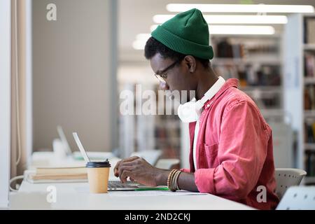Focused Black man in glasses studying online on laptop in library or classroom. Distance education.  Stock Photo