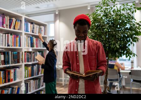 Smiling student black guy stands in library read book in background girl reach for top of bookshelf. Stock Photo
