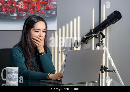 South Asian woman at desk working on laptop in home office, yawning Stock Photo