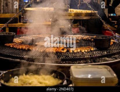 Sausages cooking on a circular hanging grill, suspended above a barbecue, with steam from a pan of onions in the foreground. Stock Photo