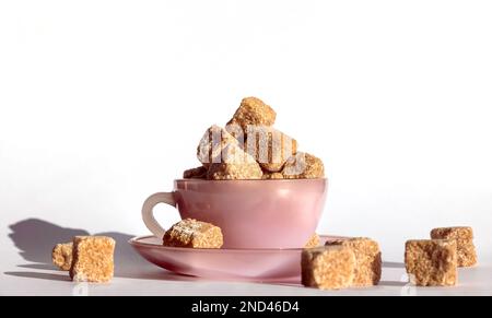 Lots of brown cane sugar cubes stacked in a teacup and saucer, on a plain white background, closeup top view, studio shot Stock Photo