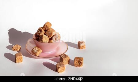 Lots of brown cane sugar cubes stacked in a teacup and saucer, on a plain white background, closeup top view Stock Photo