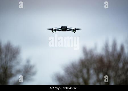 Modern quadcopter drone flying in cloudy sky framed by the blurred outlines of leafless trees below Stock Photo