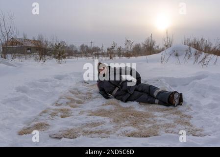 Smiling man in work clothes lying on the carpet with a long NAP in the snow on the background of building a house and snowdrifts under the setting sun Stock Photo