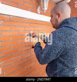 Young white man DIY with a drilling machine on the wall of his terrace. Stock Photo