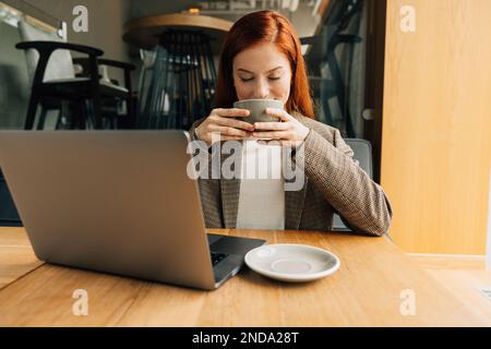 Smiling woman with ginger hair enjoying her morning coffee. Businesswoman holding a cup with closed eyes while sitting at a table. Stock Photo