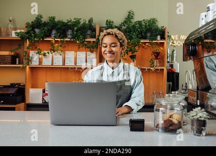 Smiling barista in apron typing on laptop at the counter. Cheerful coffee shop owner working while standing at the bar counter. Stock Photo