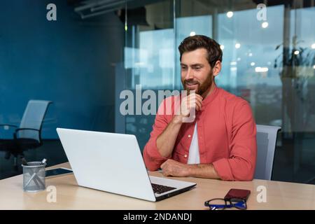 Portrait of a young male programmer, designer, freelancer in a red shirt who sits and works in the office at a laptop. He looks thoughtfully at the monitor, talks on a video call. Stock Photo