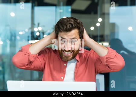 A young man sits in the office at the table, looks at the laptop monitor and rejoices. Happy and shocked holding his head, celebrating victory, success. Stock Photo