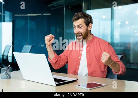 A young man sits in the office, looks at the laptop monitor and rejoices. Screams with happiness, makes victorious hand gestures. Received good news, successful deal, project, playing online games. Stock Photo