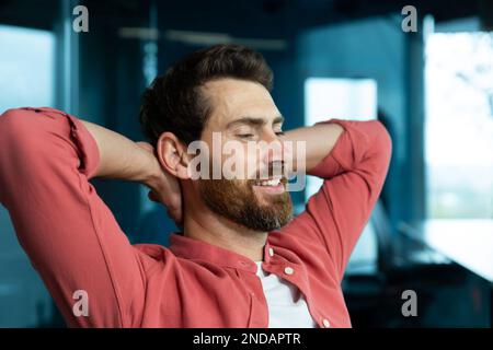 A young male programmer, a freelancer, is resting from a hard day in the office at his desk. He sits on a chair, closes his eyes, puts his hands behind his head, meditates, dreams, smiles. Stock Photo