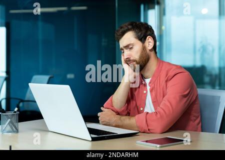 Overtime, deadline. Tired young male programmer, freelancer sits in the office and tries to work at the laptop. Bored, he sits at the table and rests his head on his hand. Stock Photo