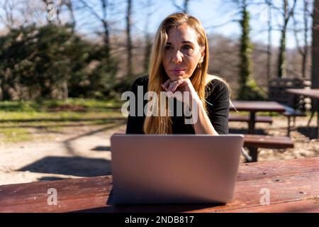 Pensive blonde haired middle aged female freelancer in black turtleneck sitting at wooden table with laptop while working remotely on project in park Stock Photo