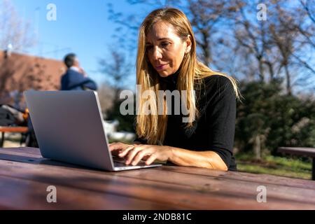 Concentrated middle aged female freelancer with blond hair wearing black turtleneck sitting at wooden table in park  Stock Photo