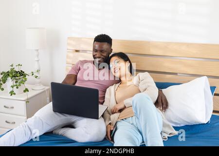 Happy diverse couple lying in bed and using laptop. Spending quality time at home, domestic life and lifestyle concept. Stock Photo
