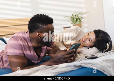 Happy diverse couple lying in bed and using smartphones. Spending quality time at home, domestic life and lifestyle concept. Stock Photo