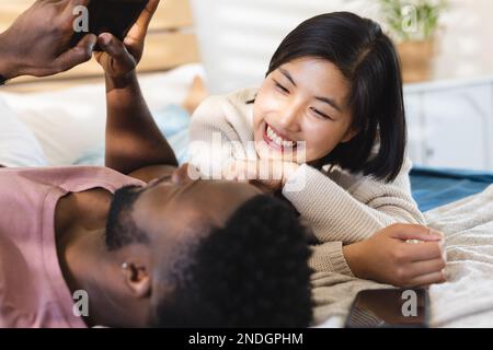 Happy diverse couple lying in bed and using smartphones. Spending quality time at home, domestic life and lifestyle concept. Stock Photo