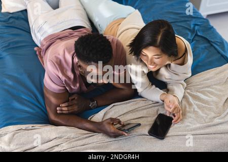 Happy diverse couple lying in bed and using smartphones. Spending quality time at home, domestic life and lifestyle concept. Stock Photo