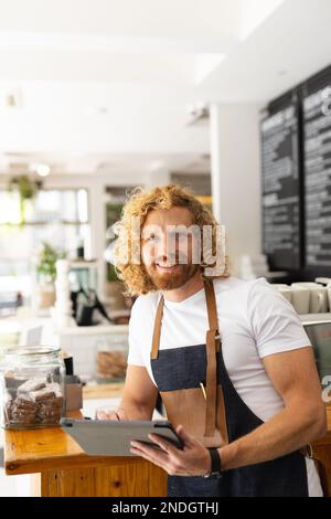 Portrait of happy caucasian male barista using tablet in cafe. Cafe, free time and city concept. Stock Photo