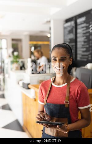 Portrait of happy biracial female barista wearing apron and using tablet in cafe. Cafe, free time and city concept. Stock Photo