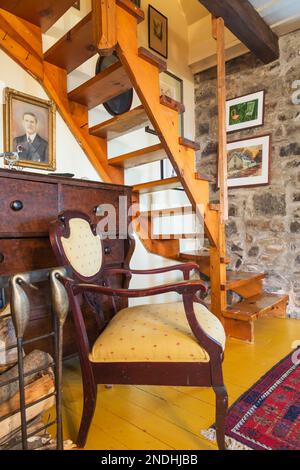 Antique upholstered armchair next to wooden dresser and twisting  staircase leading to upstairs floor in living room inside old renovated 1650 house. Stock Photo