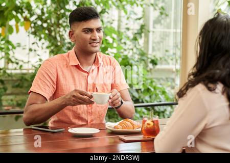 Smiling Indian man drinking coffee and talking to friends when they are sitting at cafe table Stock Photo
