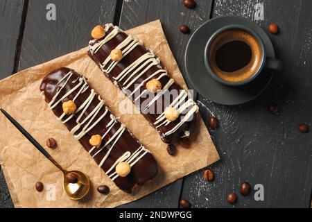 Parchment with delicious chocolate eclairs, hazelnuts and cup of coffee on black wooden background Stock Photo