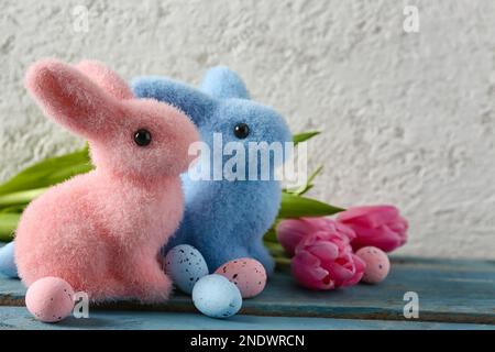 Easter bunnies, painted eggs and beautiful tulip flowers on blue wooden table near grey wall Stock Photo
