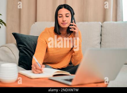 A focused mind makes things happen. a young woman using a smartphone and writing notes while working on the sofa at home. Stock Photo