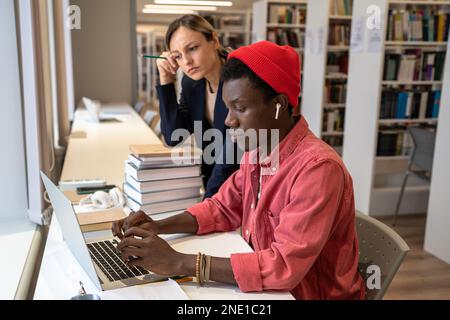 College tutor woman assists puzzled student African American guy in solve task at laptop in library. Stock Photo