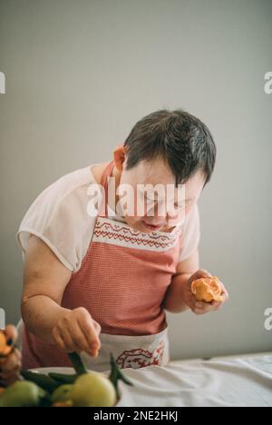 elderly woman with down syndrome is studying in the kitchen peels tangerines Stock Photo