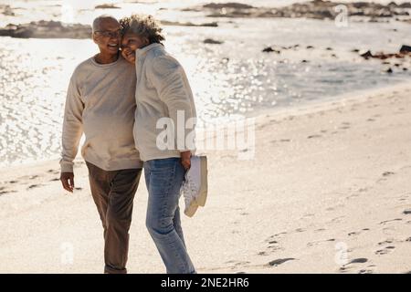 Happy elderly couple smiling and embracing each other while walking barefoot on beach sand. Romantic senior couple spending some quality time together Stock Photo