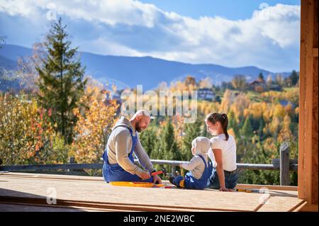 Father, mother and son building wooden frame house. Toddler boy helping his daddy, while woman looking for them on construction site. Guys wearing blue overalls. Carpentry, family concept. Stock Photo