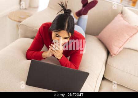 Smiling middle eastern young woman video calling through laptop while lying on sofa at home. unaltered, wireless technology, lifestyle, communication, Stock Photo