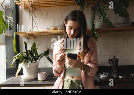 Thoughtful non-binary trans woman using smartphone in the kitchen at home. concept of gender fluidity Stock Photo