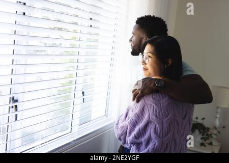 Happy diverse couple embracing and looking through window. Spending quality time at home, domestic life and lifestyle concept. Stock Photo