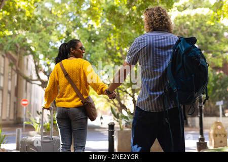 Happy diverse couple with luggage wearing sunglasses and walking in city. On the go, travel and city concept. Stock Photo