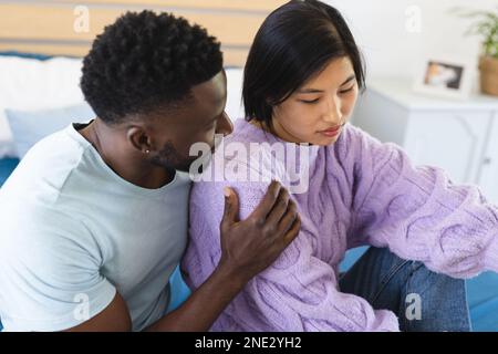 Sad diverse couple sitting on bed and embracing. Spending quality time at home, domestic life and lifestyle concept. Stock Photo