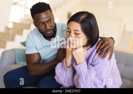Sad diverse couple sitting on sofa and embracing. Spending quality time at home, domestic life and lifestyle concept. Stock Photo