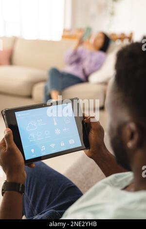 African american man sitting on sofa in living room and using tablet with smart home interface. Spending quality time at home, domestic life and lifes Stock Photo