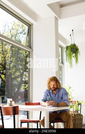 Happy caucasian man sitting at table and using smartphone in cafe. Cafe, free time and city concept. Stock Photo