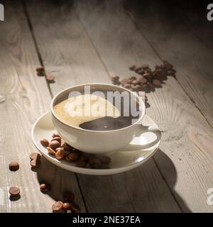 White cup of hot coffee with steam on a wooden table backlit. Stock Photo