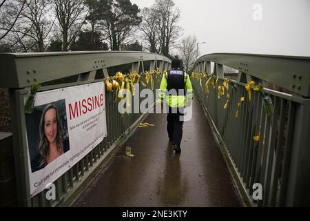 A police officer walks past a missing person appeal poster for Nicola Bulley and yellow ribbons and messages of hope tied to a bridge over the River Wyre in St Michael's on Wyre, Lancashire, as police continue their search for Ms Bulley, 45, who vanished on January 27 while walking her springer spaniel Willow shortly after dropping her daughters, aged six and nine, at school. Picture date: Thursday February 16, 2023. Stock Photo