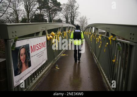 A police officer walks past a missing person appeal poster for Nicola Bulley and yellow ribbons and messages of hope tied to a bridge over the River Wyre in St Michael's on Wyre, Lancashire, as police continue their search for Ms Bulley, 45, who vanished on January 27 while walking her springer spaniel Willow shortly after dropping her daughters, aged six and nine, at school. Picture date: Thursday February 16, 2023. Stock Photo