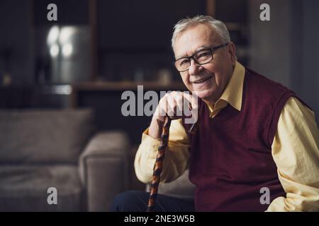 Happy senior man is sitting on bed in his home. Portrait of senior man with walking cane. Stock Photo