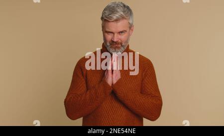 Sneaky cunning middle-aged man with tricky face gesticulating and scheming evil plan, thinking over devious villain idea, cheats, revenge, jokes, pranks. Senior guy isolated on beige studio background Stock Photo