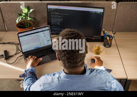 Black Man Writing Lines of Code On Desktop PC With Two Monitors and a Laptop Aside in Stylish Office. Professional Male Developer Programming Stock Photo