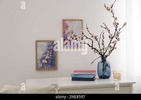 Flowering tree twigs in glass vase, burning candle and books on table near white wall at home Stock Photo