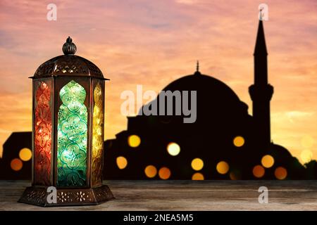 Decorative Arabic lantern on wooden surface and silhouette of mosque at sunset on background, space for text Stock Photo