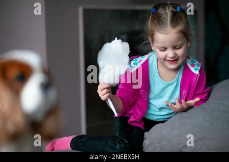 Portrait of smiling little girl holding candyfloss, looking at sticky fingers hand, sitting on sofa near dog at home. Stock Photo